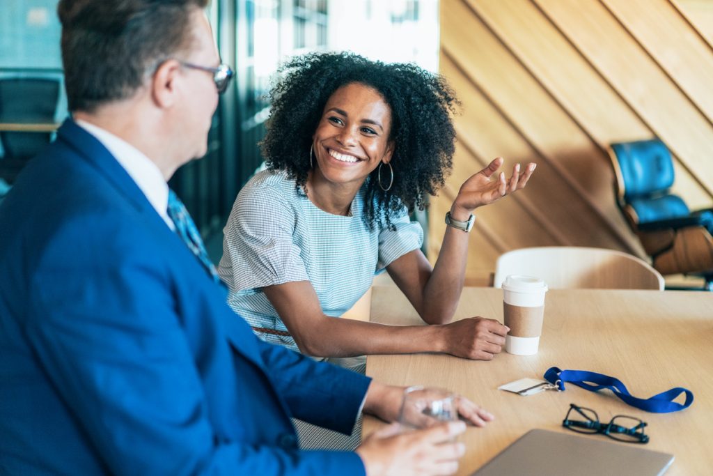Two colleagues sit at a wooden table in a modern office, engaged in a friendly conversation. One person is smiling and gesturing with a hand, holding a coffee cup in front of them, while the other listens attentively with a glass of water in hand. A lanyard, glasses, and a laptop rest on the table.