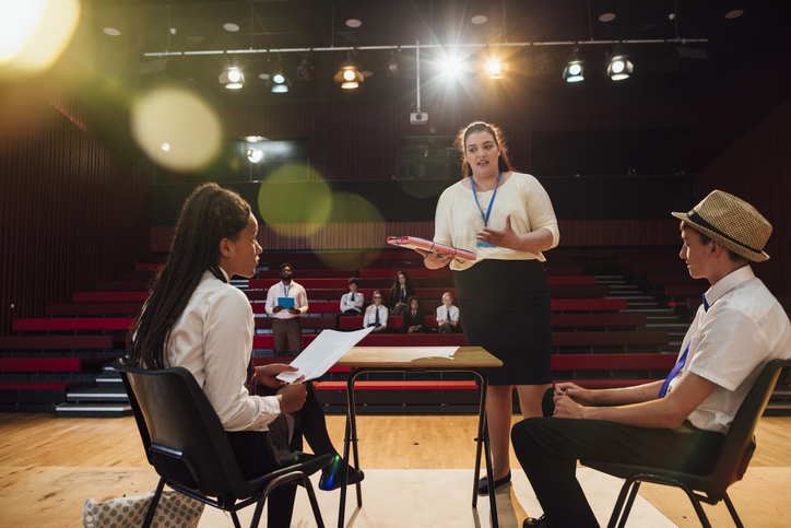 An over the shoulder view of two students who are taking notes on performace and how to improve in rehearsal. They are being watched by the rest of the drama club in the theatre.