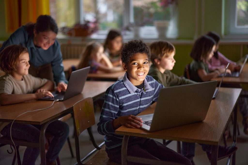 A cheerful classroom scene with elementary-aged students working on laptops. A smiling boy in the foreground looks directly at the camera, while other students and a teacher engage in learning activities in the background. The classroom is softly lit, with large windows and warm yellow curtains.