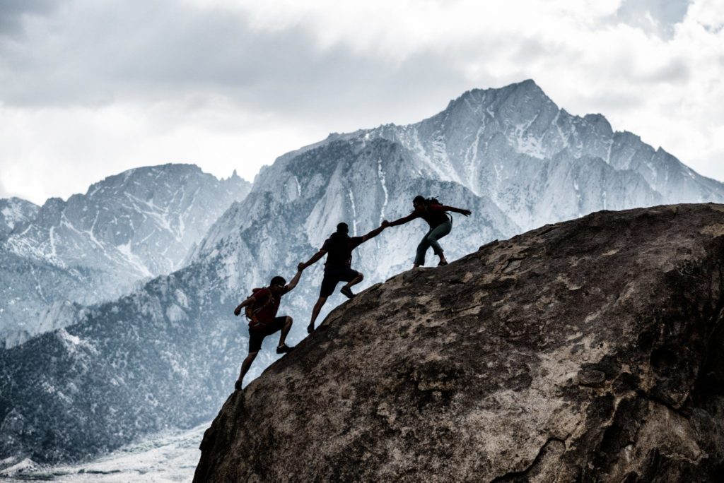3 men climbing a mountain. All three are helping eachother climb.